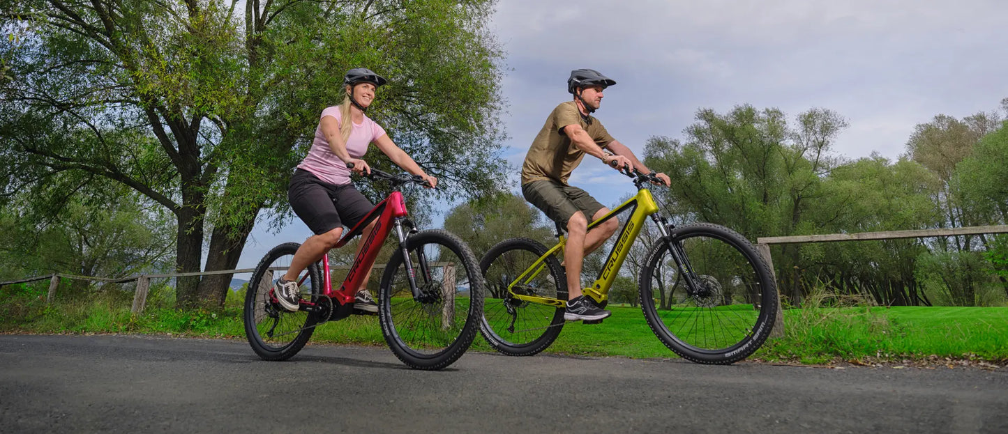 Crussis ONE-Guera 10.10 Women’s Mountain E-Bike. Two people riding electric bikes on a road with trees and grass in the background.